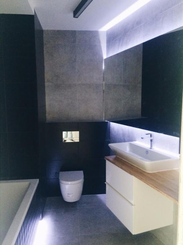 Nowoczesna łazienka w szaro-czarnych tonacjach z podświetleniem LED: umywalka nablatowa na białej szafce z drewnianym blatem, wisząca toaleta i minimalistyczny design.