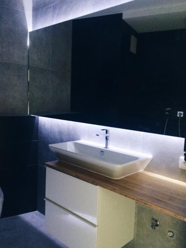 Nowoczesna łazienka z podświetlanym lustrem i umywalką nablatową na szafce z drewnianym blatem, szare płytki na ścianach.