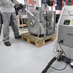 Pracownik w kombinezonie obsługuje urządzenie do czyszczenia form wtryskowych, umieszczonych na palecie, w hali produkcyjnej.