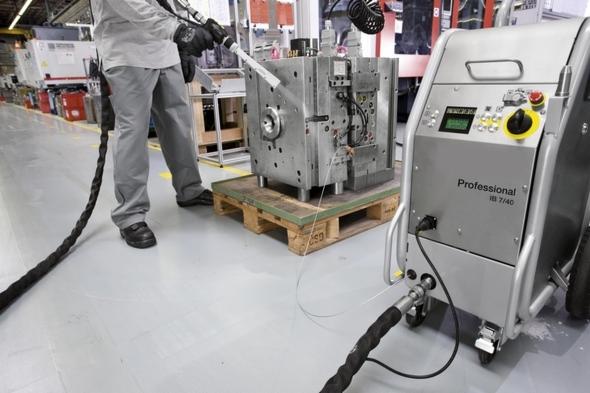 Pracownik w kombinezonie obsługuje urządzenie do czyszczenia form wtryskowych, umieszczonych na palecie, w hali produkcyjnej.