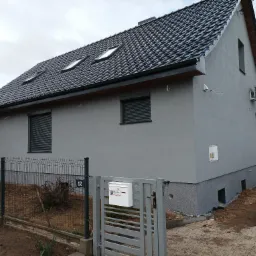 Nowoczesny dom jednorodzinny z szarą elewacją, ciemną dachówką i oknami z roletami zewnętrznymi, widoczny z perspektywy ogrodu z metalowym ogrodzeniem i skrzynką pocztową z naklejką 'Uwaga kot'.