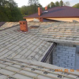 Widok z góry na konstrukcję dachu z łatami i folią, częściowo pokrytą dachówką, z kominem z czerwonej cegły i widocznym fragmentem sąsiedniego budynku.