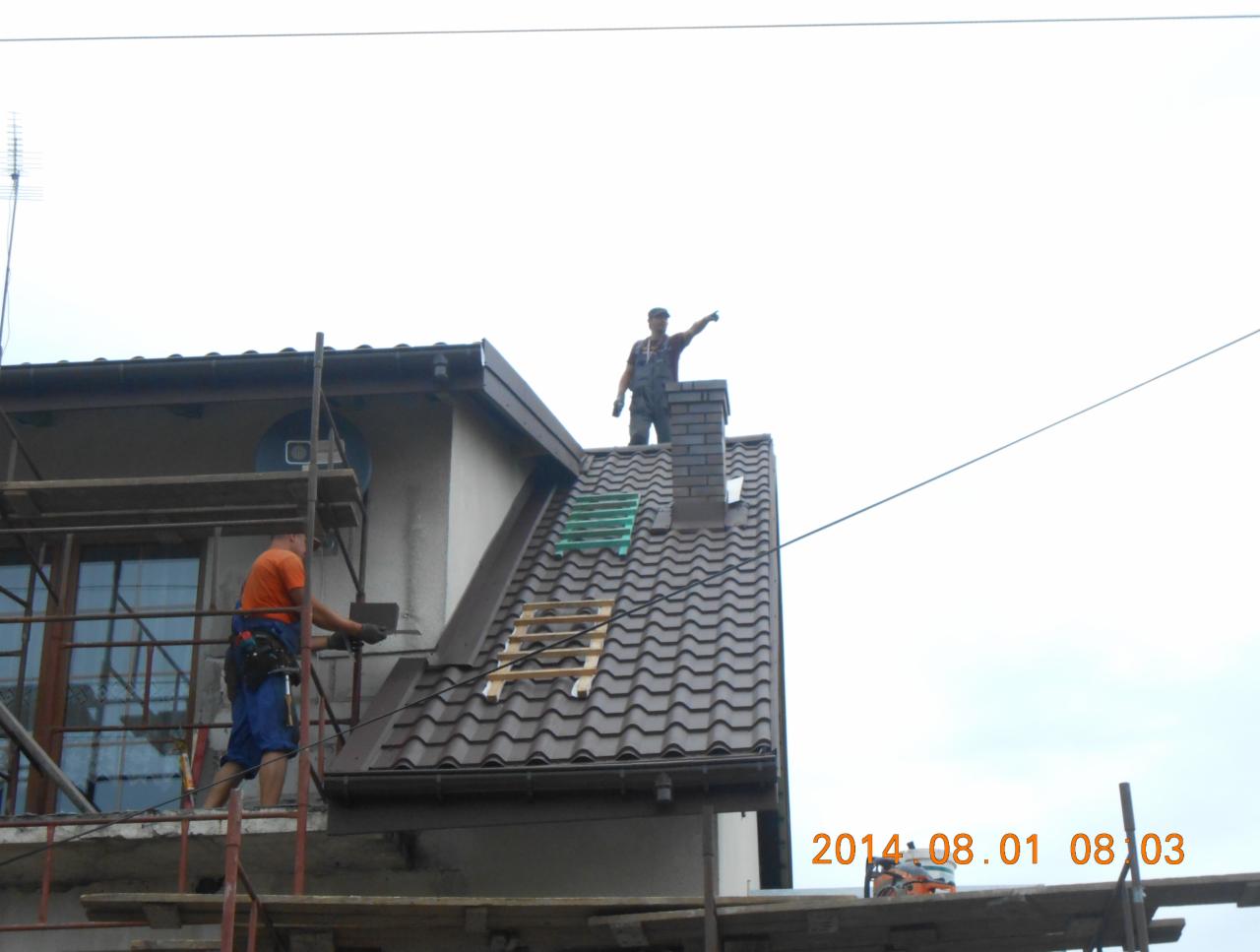 Dekarze pracują na dachu domu jednorodzinnego z brązową dachówką, jeden na rusztowaniu układa dachówkę, drugi stoi na kominie i wskazuje palcem.