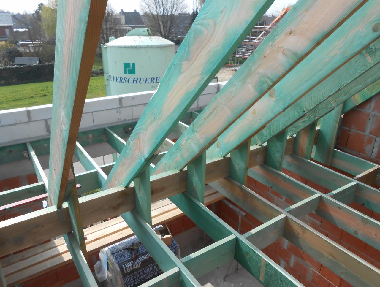 Konstrukcja drewniana więźby dachowej z impregnacją na zielono, widok z góry na ceglany mur i częściowo ułożone deski, w tle zbiornik na wodę.