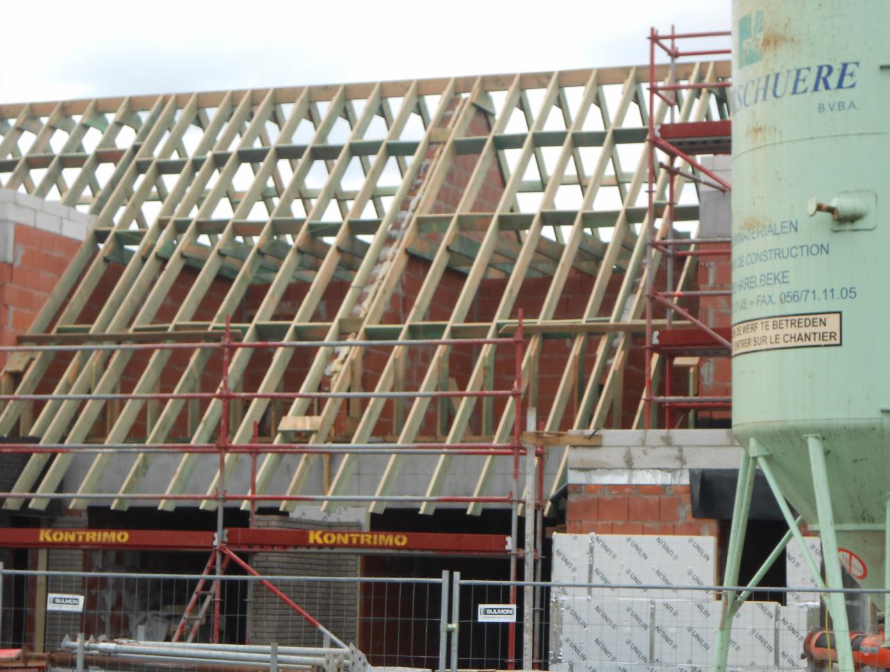 Konstrukcja więźby dachowej z czerwonej cegły w trakcie budowy, widoczne rusztowania i silos na zaprawę, zabezpieczenia budowy w tle.