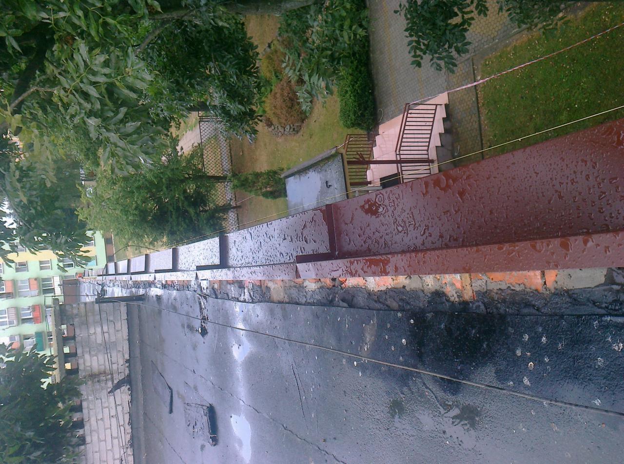 Widok z góry na płaski dach pokryty papą, z widocznymi obróbkami blacharskimi i fragmentem budynku mieszkalnego w tle, po deszczu.