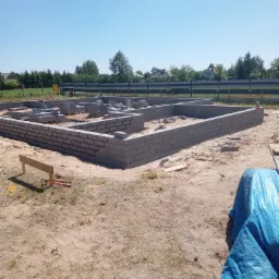 Wznoszenie fundamentów z bloczków betonowych na piaszczystym gruncie, widoczne narzędzia i niebieska plandeka w rogu kadru, w tle zieleń i błękitne niebo.