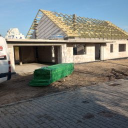 Mar bud - Budowa domu z pustaków: widoczny szkielet dachu z drewnianych belek, paleta zielonych desek na pierwszym planie, biały samochód dostawczy z boku.