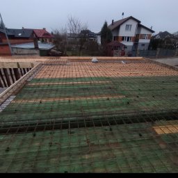 Mar bud - Zbrojenie stropu na deskowaniu, widok z góry na siatkę z prętów stalowych, drewniane podpory i folię izolacyjną, w tle zabudowa jednorodzinna w pochmurny dzień.