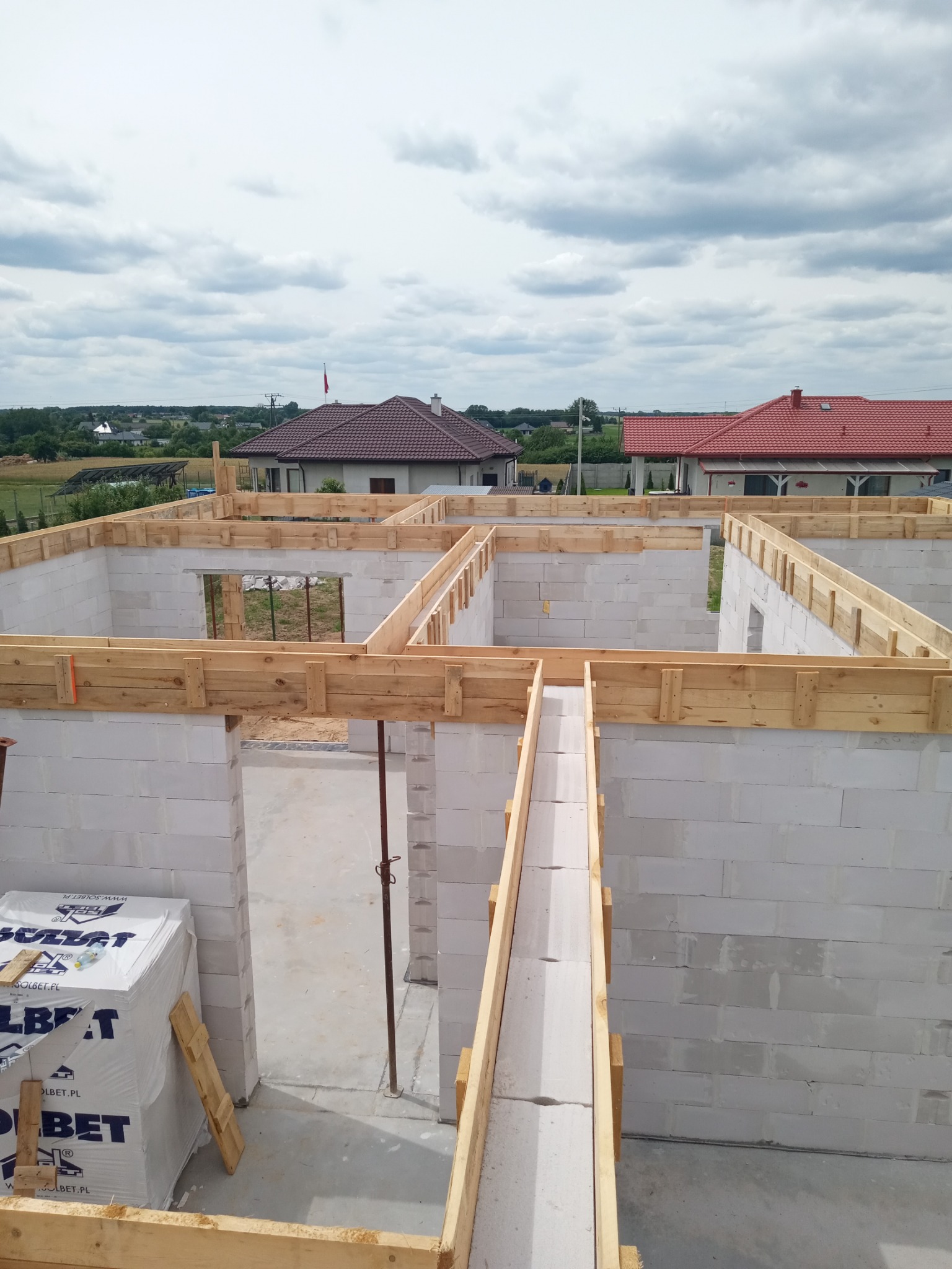 Budowa domu z bloczków betonu komórkowego, widoczne szalunki drewniane i paleta bloczków Solbet, w tle zabudowa jednorodzinna i pochmurne niebo.