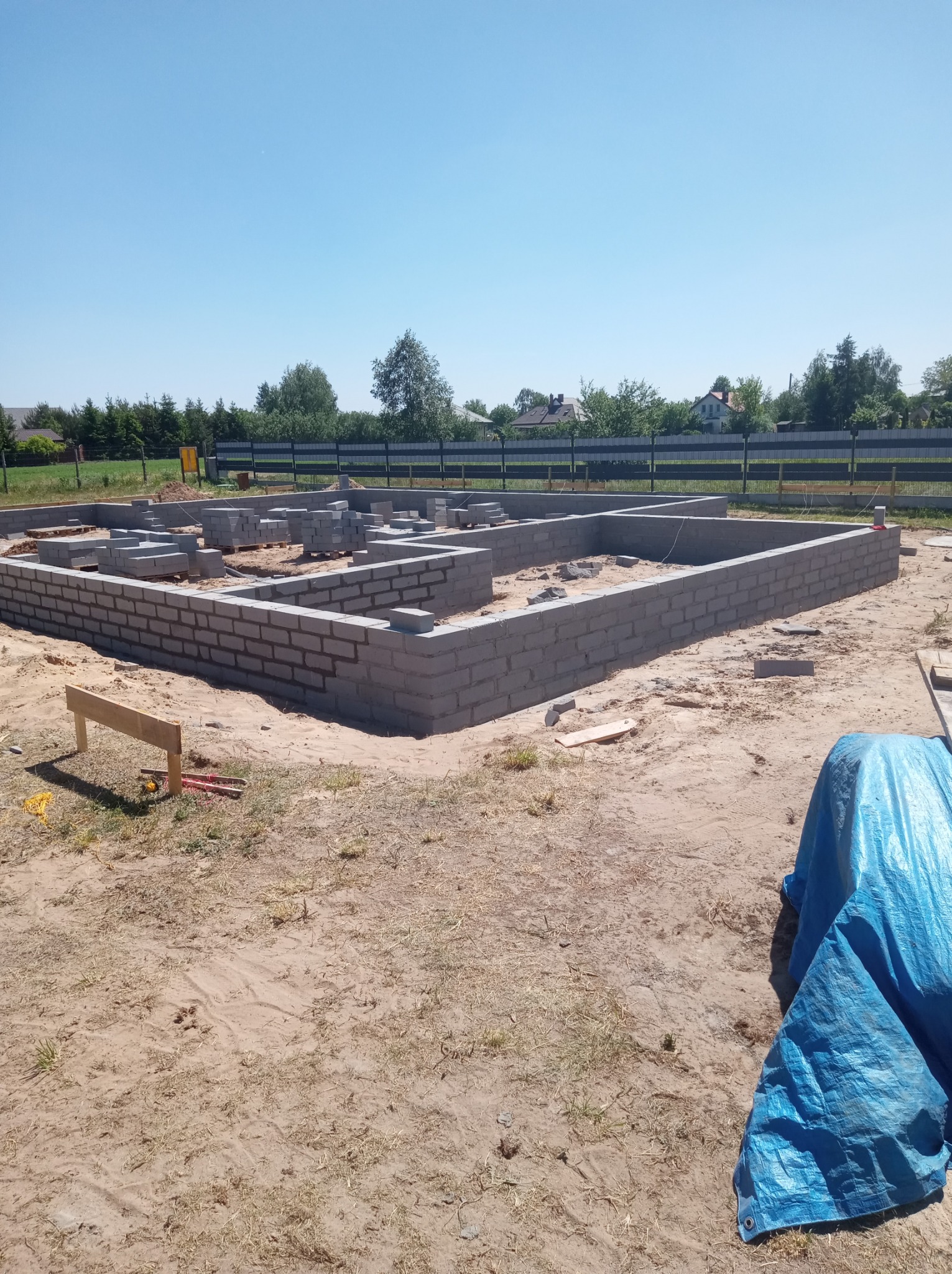 Wznoszenie fundamentów z bloczków betonowych na piaszczystym gruncie, widoczne narzędzia i niebieska plandeka w rogu kadru, w tle zieleń i błękitne niebo.