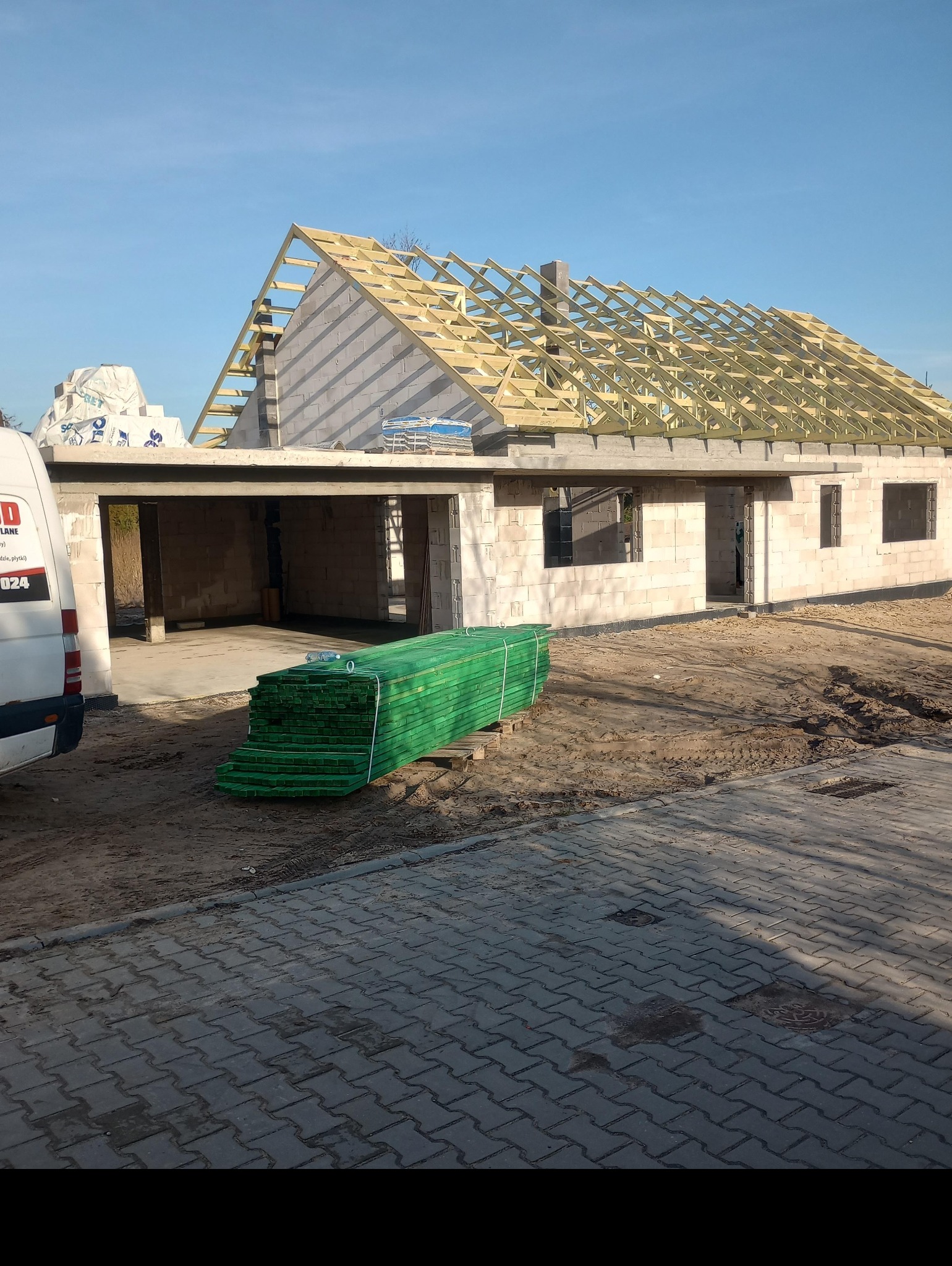 Budowa domu z pustaków: widoczny szkielet dachu z drewnianych belek, paleta zielonych desek na pierwszym planie, biały samochód dostawczy z boku.