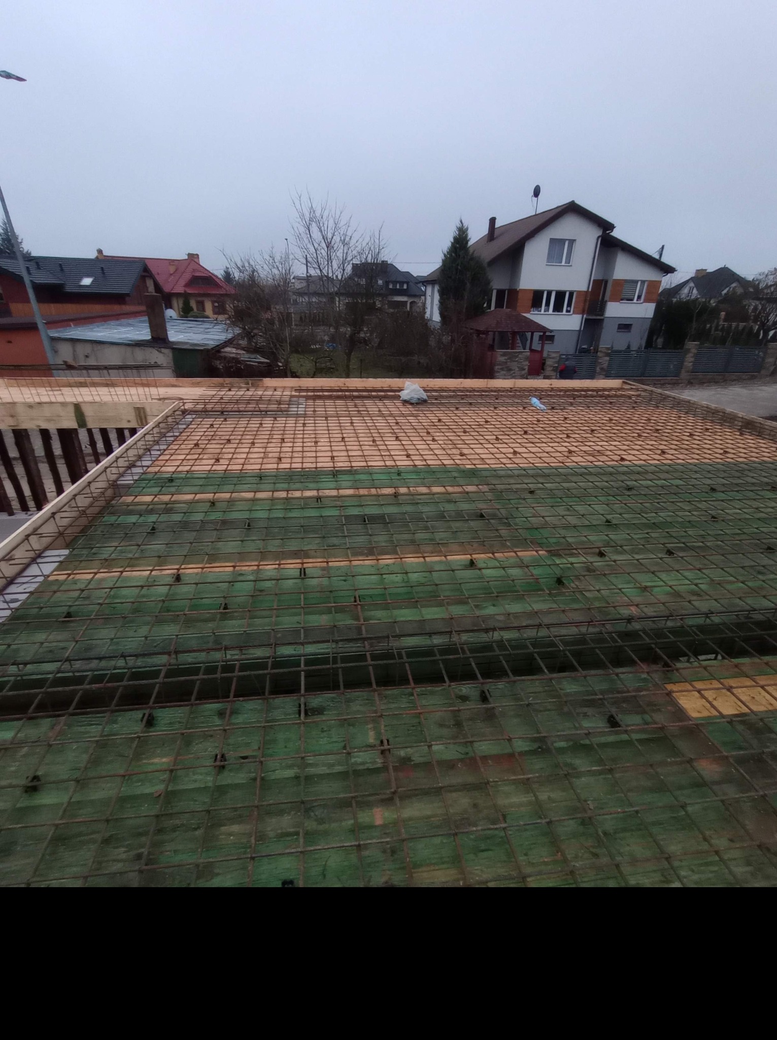 Zbrojenie stropu na deskowaniu, widok z góry na siatkę z prętów stalowych, drewniane podpory i folię izolacyjną, w tle zabudowa jednorodzinna w pochmurny dzień.