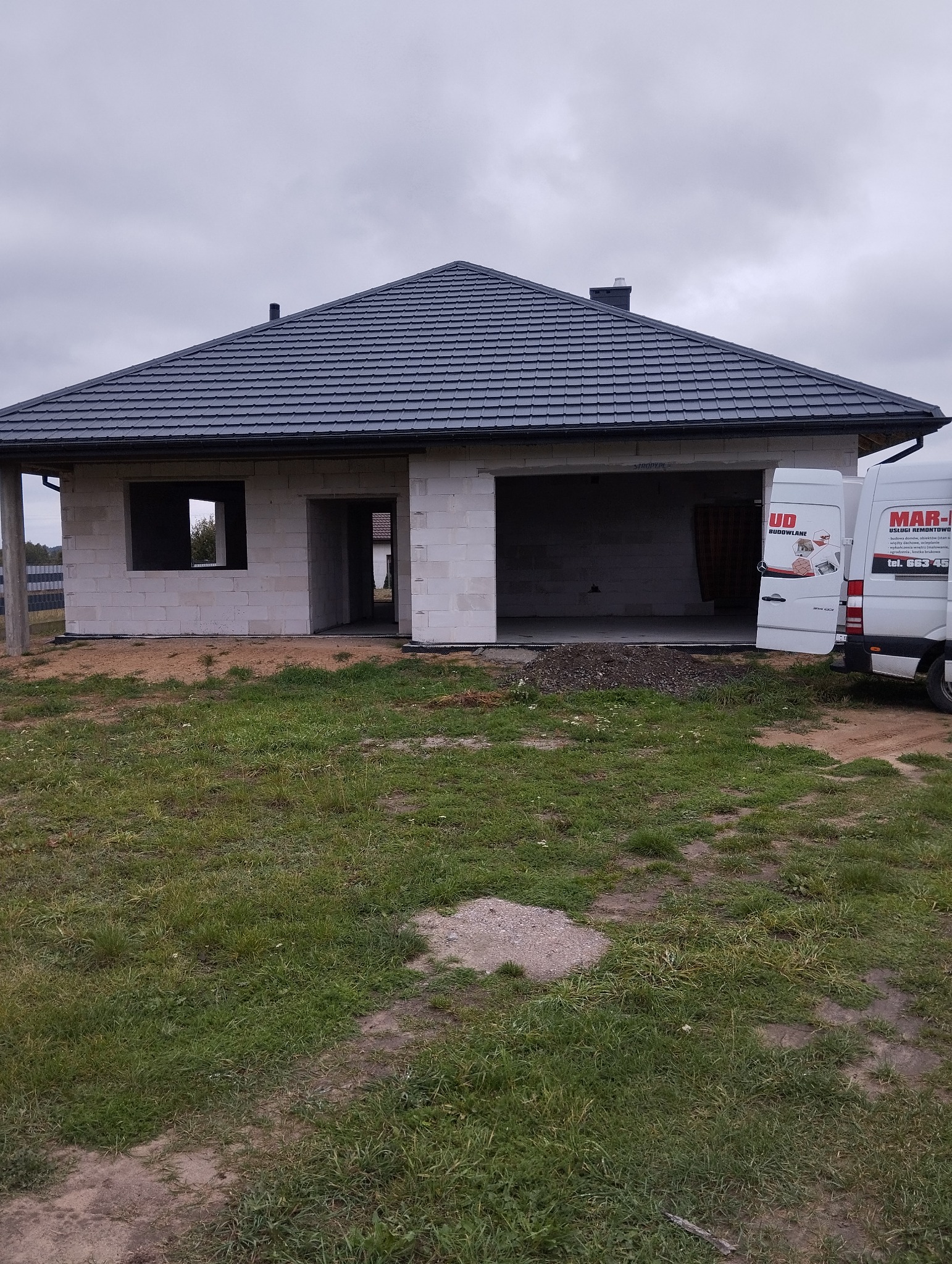 Surowy stan domu z szarej dachówki, widoczne białe bloczki, otwarte okna i drzwi, oraz firmowe busy z reklamami usług budowlanych w tle.