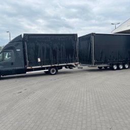 Arkadiusz Nowak - Czarny bus z przyczepą plandekową na betonowym placu, widok z boku. Na przyczepie widoczne oznaczenia ostrzegawcze. W tle słup z czerwonymi pasami i zadaszenie.