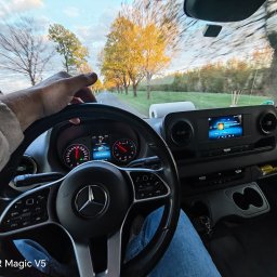 Arkadiusz Nowak - Wnętrze busa Mercedes z perspektywy kierowcy podczas jazdy po drodze otoczonej drzewami. Widoczna deska rozdzielcza, kierownica i ręka na kierownicy.