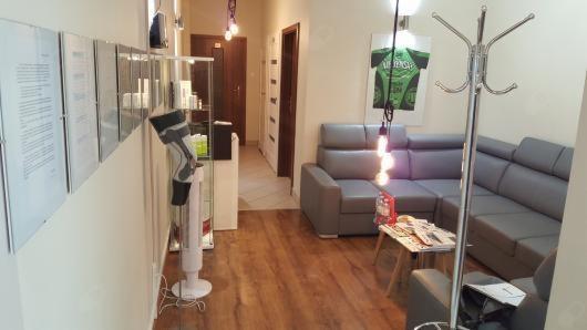 Wnętrze poczekalni: szary narożnik, stolik z gazetami, wieszak na ubrania, grafika na ścianie, dokumenty w ramkach na ścianie, podłoga imitująca drewno.