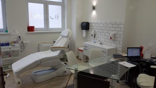 Jasne wnętrze gabinetu zabiegowego z białym fotelem kosmetycznym, umywalką z kafelkami w stylu metra i szklanym biurkiem z laptopem.