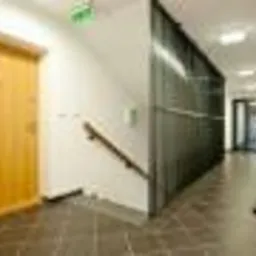 Korytarz z drewnianymi drzwiami i schodami z balustradą. Podłoga wyłożona ciemnymi płytkami. Widoczna metalowa konstrukcja. Jasne, neutralne oświetlenie.