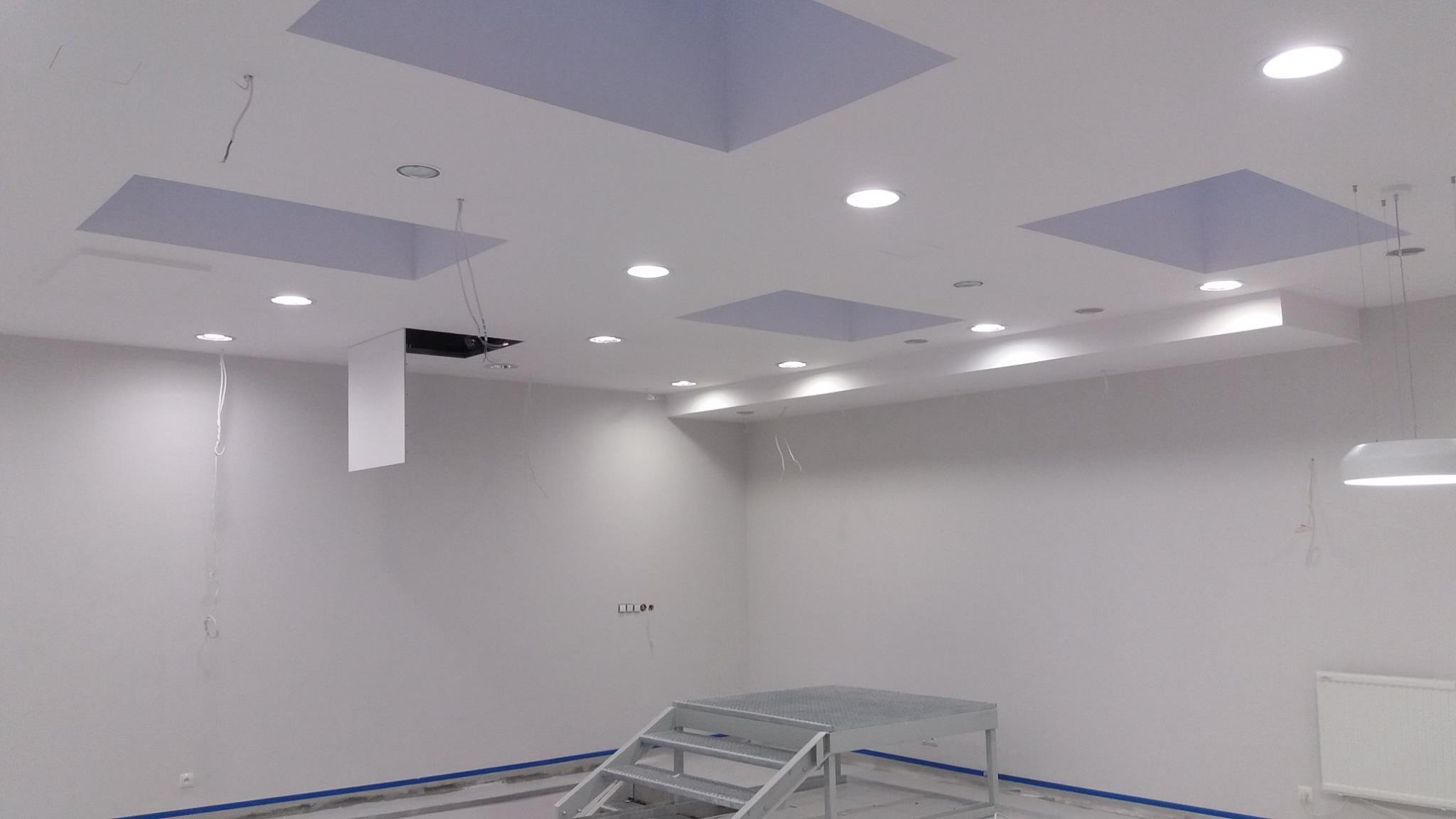 Wnętrze w trakcie remontu, widoczne przygotowane otwory w suficie pod podwieszane panele, metalowy podest roboczy i zabezpieczona podłoga.