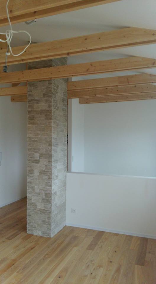 Wnętrze w trakcie remontu: drewniane belki stropowe, kolumna obłożona dekoracyjnym kamieniem, parkiet na podłodze, białe ściany, widoczne przewody elektryczne.