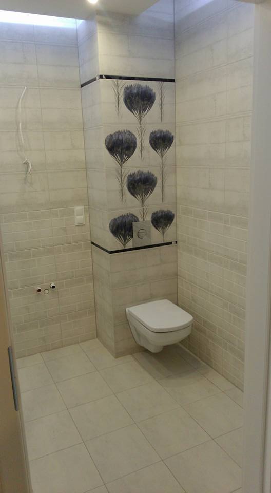 Ściana w łazience wyłożona jasnymi płytkami z motywem kwiatowym nad zabudowaną toaletą podtynkową, widoczne przyłącza wodne i fragment instalacji elektrycznej.