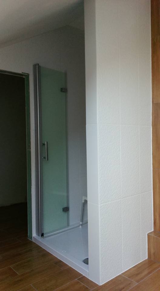 Kabina prysznicowa z szklanymi drzwiami w łazience wykończonej białymi, strukturalnymi płytkami i drewnopodobną podłogą.