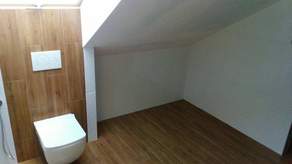 Nowoczesna łazienka na poddaszu z drewnopodobnymi płytkami na podłodze i ścianie, wisząca toaleta z białym przyciskiem spłukującym, białe ściany pod skosem.