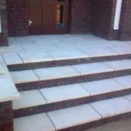 Czterostopniowe schody zewnętrzne prowadzące do drzwi wejściowych, wyłożone jasnymi, prostokątnymi płytami z obramowaniem z ciemnej cegły, widoczne fragmenty ściany budynku z cegły i białe elementy...