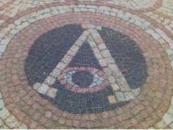 Kostka brukowa ułożona w okrągły wzór z centralnym motywem litery 'A' i stylizowanym okiem wewnątrz, wykonana z różnokolorowych kostek tworzących koncentryczne okręgi.