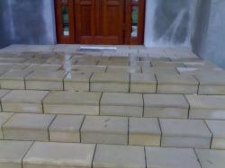 Zewnętrzne schody z jasnych, kwadratowych płyt kamiennych prowadzące do drewnianych drzwi wejściowych. Widoczne ślady wilgoci na powierzchni kamienia.