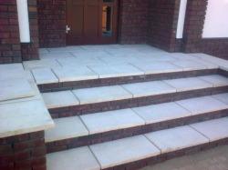 Czterostopniowe schody zewnętrzne prowadzące do drzwi wejściowych, wyłożone jasnymi, prostokątnymi płytami z obramowaniem z ciemnej cegły, widoczne fragmenty ściany budynku z cegły i białe elementy...