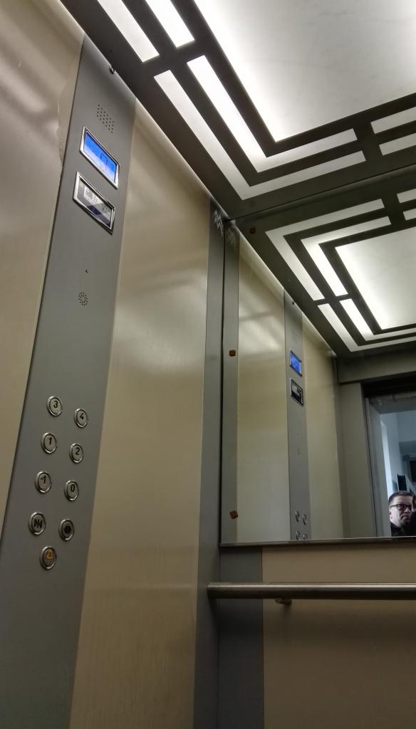 Wnętrze windy z panelem sterowania po lewej, odbicie osoby w lustrze na ścianie windy i geometryczny wzór na suficie. Metalowe wykończenie.