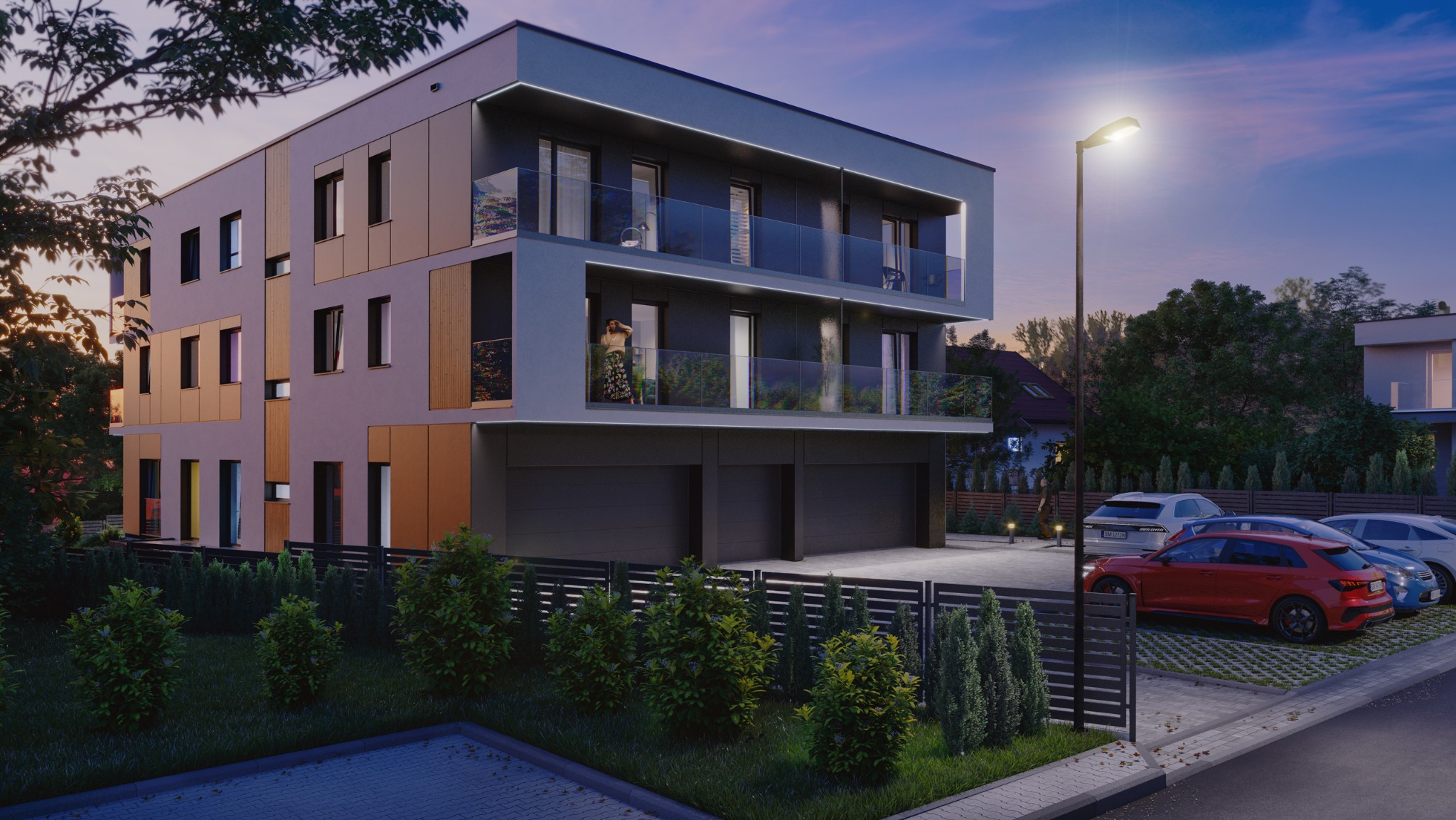 Nowoczesny budynek wielorodzinny z balkonami i garażami, oświetlony latarnią uliczną, z zaparkowanymi samochodami i zielenią w tle, widziany o zmierzchu.