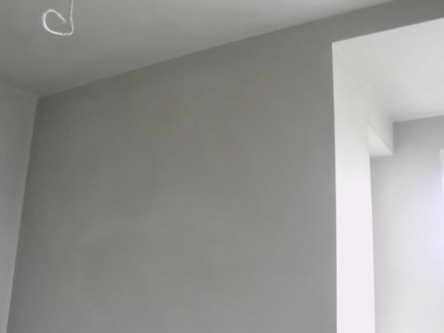 Świeżo położony, gładki, szary tynk na ścianie wewnętrznej. W rogu widoczny fragment okna z białym ościeżem. U góry zwisający kabel.