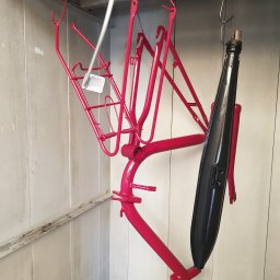 Soda Blaster - Czerwona rama roweru i czarny błotnik zawieszone na hakach w kabinie lakierniczej, po świeżym malowaniu.