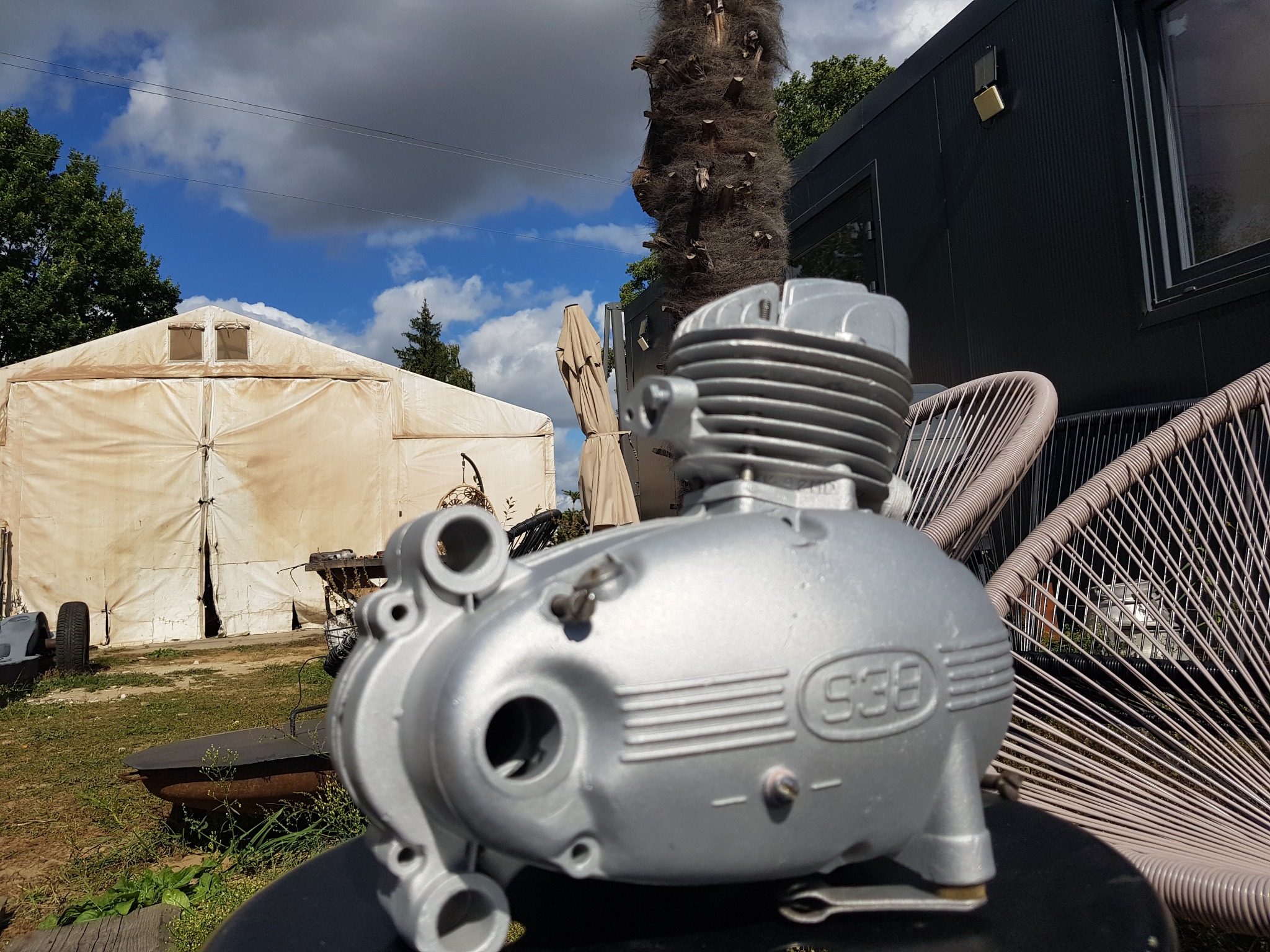 Srebrny silnik motocyklowy S38 po renowacji, umieszczony na tle ogrodu z białym namiotem, palmą i nowoczesnym budynkiem.