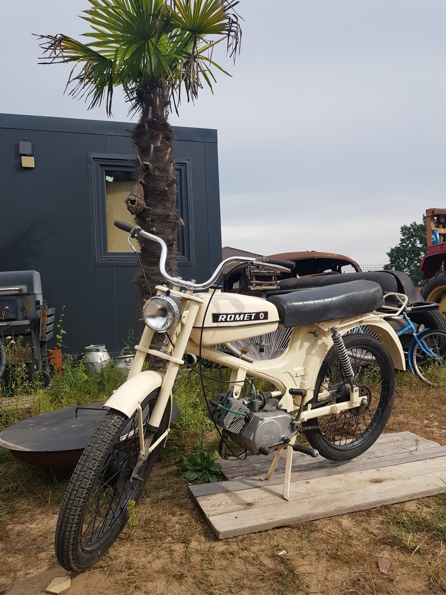 Kremowy motorower Romet na drewnianej platformie, na tle palmy i garażu blaszanego, obok starego samochodu i roweru.