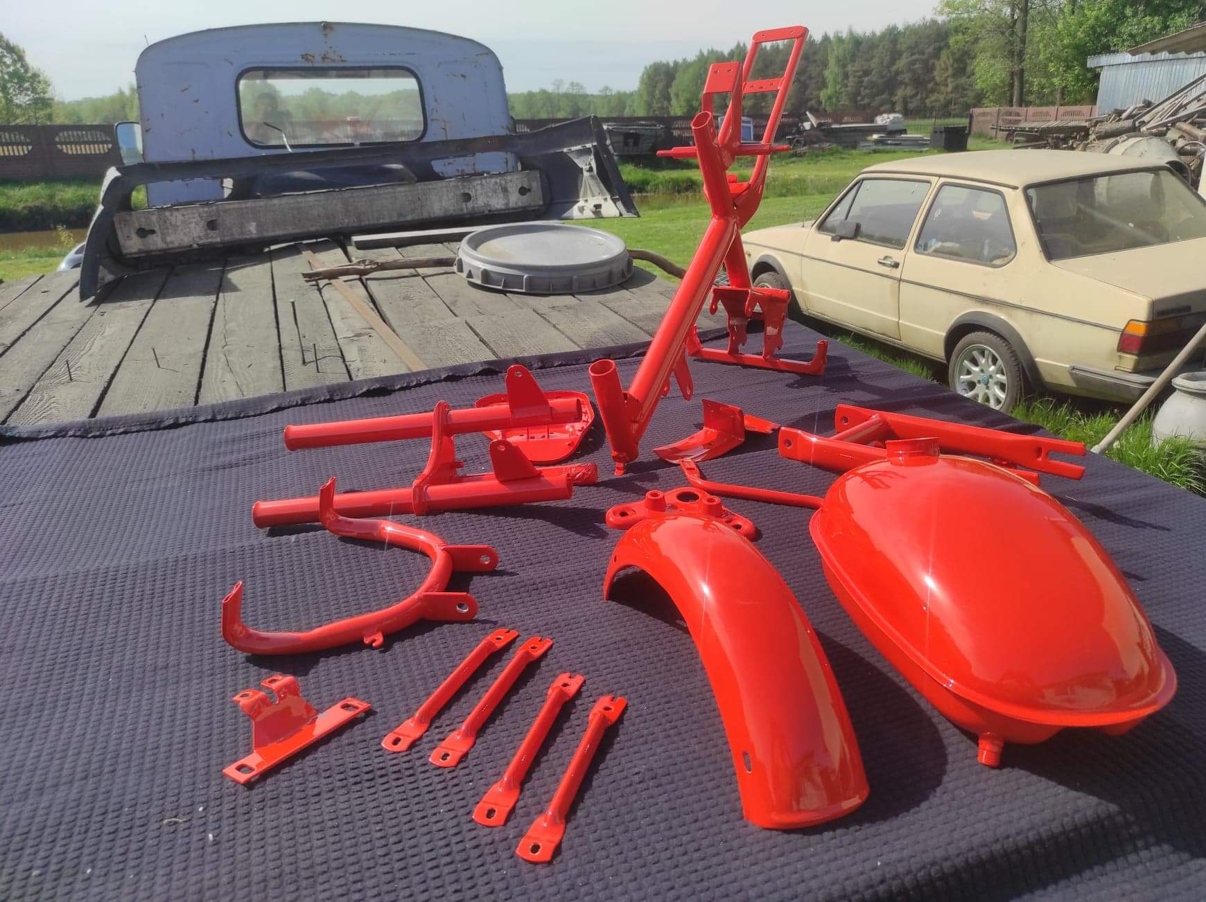 Części metalowe pomalowane na czerwono, leżące na ciemnej tkaninie, w tle samochód i platforma ciężarówki.