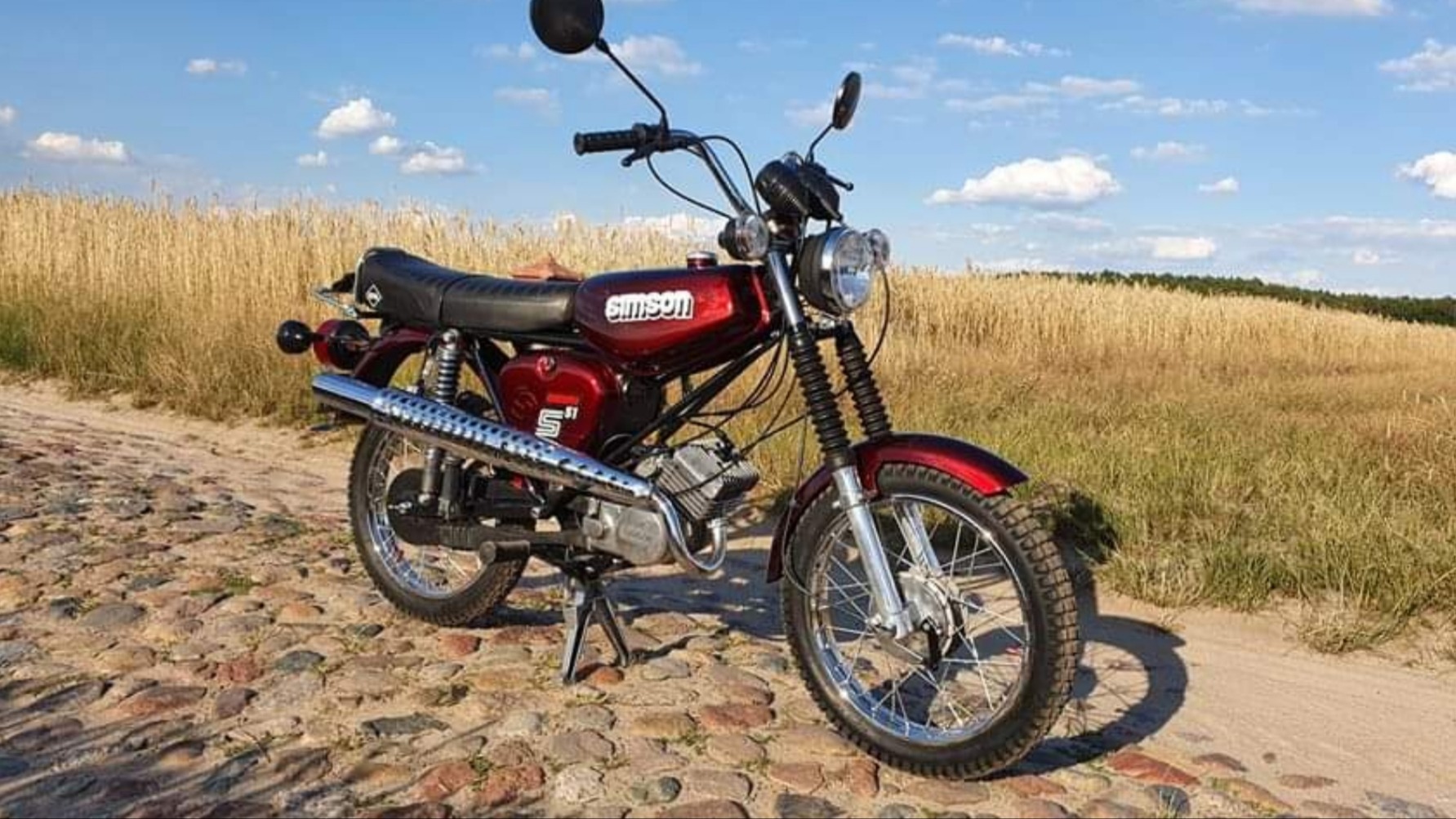 Czerwony motocykl Simson S51 na tle pola zboża i błękitnego nieba, stojący na kamiennej drodze.