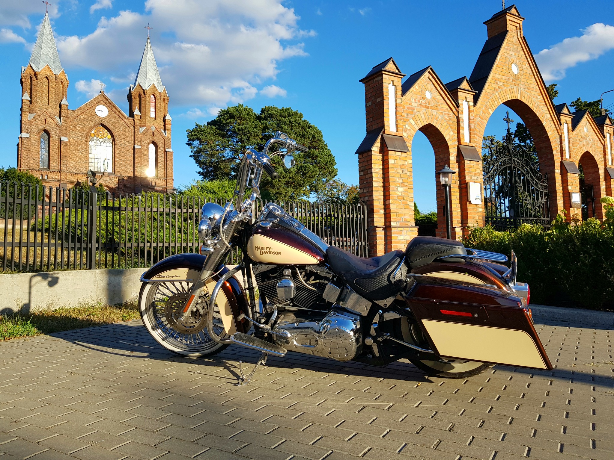 Motocykl Harley-Davidson Deluxe w kolorze bordowo-kremowym na kostce brukowej, na tle ceglanego kościoła z dwiema wieżami i ceglanej bramy z kutą kratą.