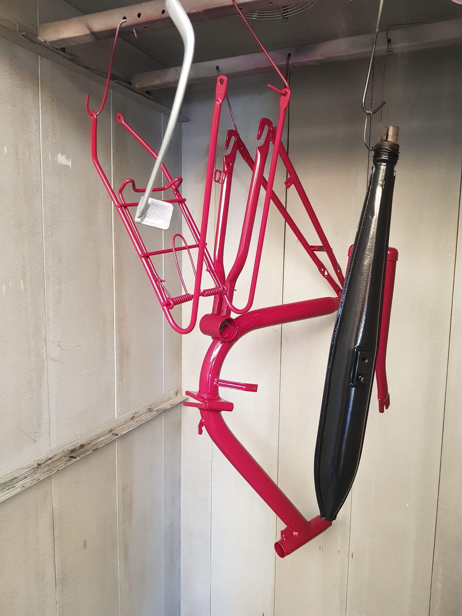 Czerwona rama roweru i czarny błotnik zawieszone na hakach w kabinie lakierniczej, po świeżym malowaniu.