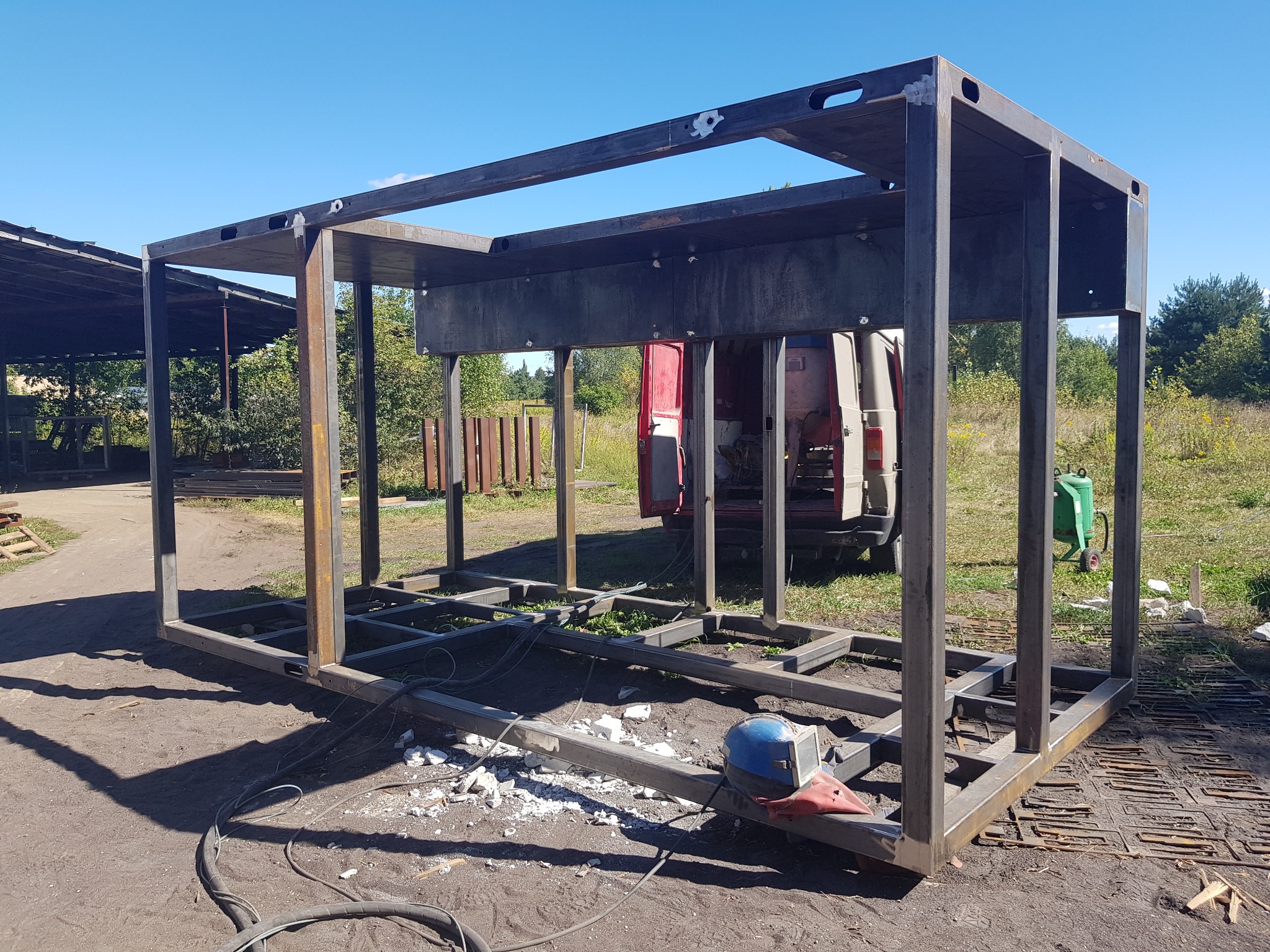 Konstrukcja szkieletowa z metalowych profili, prawdopodobnie pod kontener, spawana na zewnątrz, widoczne ślady spawania, kask spawalniczy i przewody spawalnicze, w tle częściowo widoczny samochód...