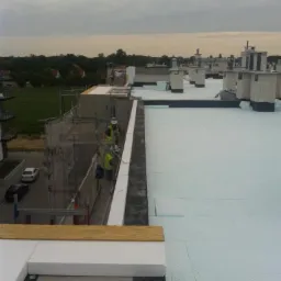 Prace na dachu: robotnicy w kamizelkach układają izolację z białego styropianu. Widok z góry na częściowo ocieplony dach budynku, w tle zabudowa miejska i zieleń.