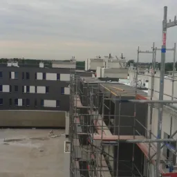 Budynek w trakcie ocieplania, widoczne rusztowania i płyty izolacyjne. Szary budynek z regularnymi oknami w tle, pochmurne niebo. Prace na wysokości.