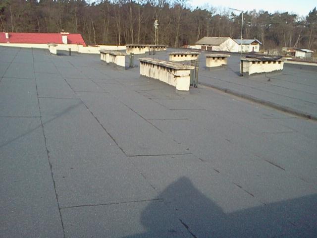 Szary, płaski dach pokryty papą z widocznymi kominami wentylacyjnymi i budynkami w tle. Widoczne łączenia papy i drobne uszkodzenia powierzchni.