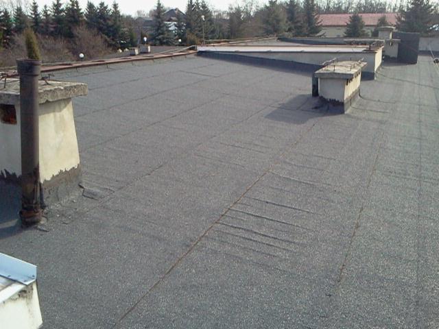 Płaski dach pokryty papą z kominami i elementami wentylacyjnymi. Widoczne nierówności i potencjalne miejsca przecieków. Tło stanowią drzewa i budynki.