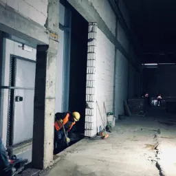 Pracownik w kasku i kamizelce odblaskowej wyrównuje podłoże wewnątrz budowanego obiektu z białych pustaków, widoczne szczeliny w posadzce i przygotowane otwory drzwiowe.