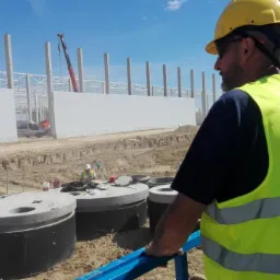 Pracownik w żółtej kamizelce z logo firmy na tle budowy hali z płyt warstwowych, widoczny dźwig i wykop z betonowymi kręgami.