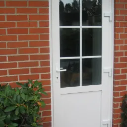 Białe drzwi zewnętrzne z sześcioma szybkami, zamontowane w ścianie z czerwonej cegły, z widocznymi roślinami po bokach.